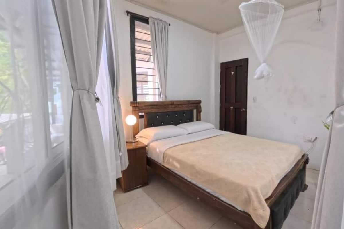 A bedroom with a bed, bedside table, lamp, window with curtains, and a door.