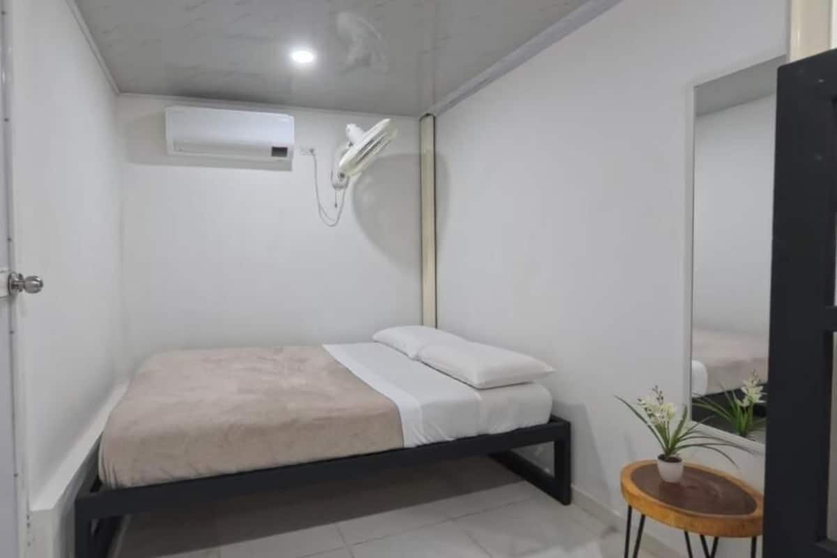 A small, modern bedroom with a bed, a small table with a plant, a mirror, and a wall-mounted air conditioning unit.