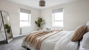 A bedroom with a bed, two windows, a radiator, a plant, and a mirror. - Modern 4BR Home 10 Beds Family-Friendly, Aylesbury (Bierton)