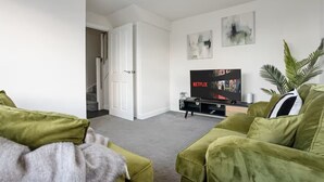 A living room with green sofas, a TV on a stand, and abstract art on the wall. - Modern 4BR Home 10 Beds Family-Friendly, Aylesbury (Bierton)
