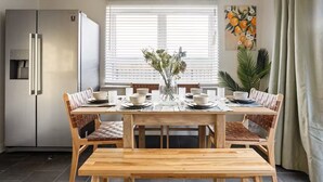 A dining area with a wooden table set for four, surrounded by cushioned chairs, a refrigerator, and a window with blinds. - Modern 4BR Home 10 Beds Family-Friendly, Aylesbury (Bierton)
