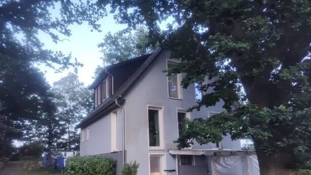 Ein zweistöckiges Haus mit geneigtem Dach, einem sichtbaren Fenster und einem Baum im Vordergrund.