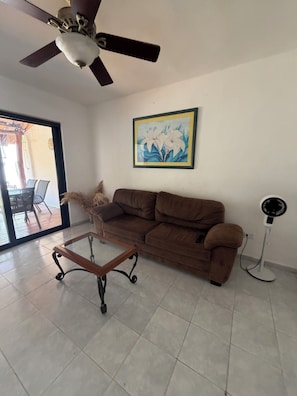 A living room with a brown sofa, a glass coffee table, a ceiling fan, a framed picture, and a standing fan. - Beautiful house next to the beach (Cancún)