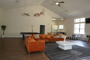 Un vaste espace de vie comprenant une table de billard, des canapés orange, une table basse blanche, des ventilateurs de plafond et de grandes fenêtres.