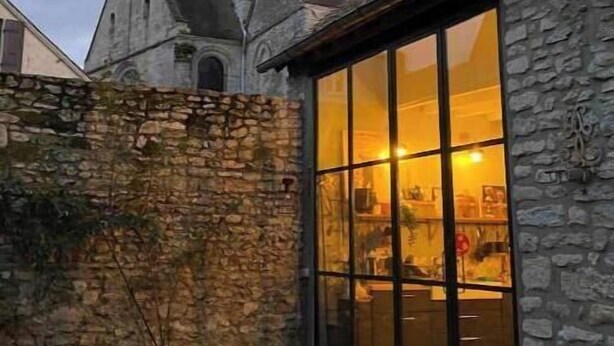 A stone building with a large glass window, a stone wall, and a tower with a clock. - Charming stone house in the heart of the Vexin, enclosed garden and view of the bell tower (Vaudancourt)