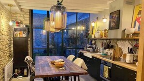 A modern kitchen with a dining table, chairs, a hanging light fixture, and a view through large windows. - Charming stone house in the heart of the Vexin, enclosed garden and view of the bell tower (Vaudancourt)
