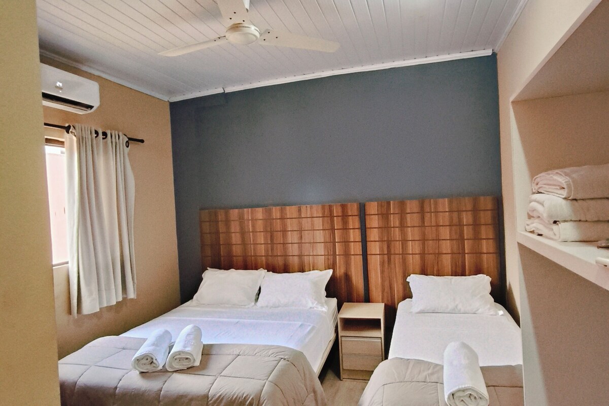 A hotel room with two beds, a ceiling fan, an air conditioning unit, and a window with curtains.