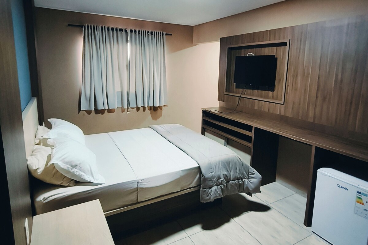 A compact room with a bed, a television mounted on the wall, a small refrigerator, and a curtain.