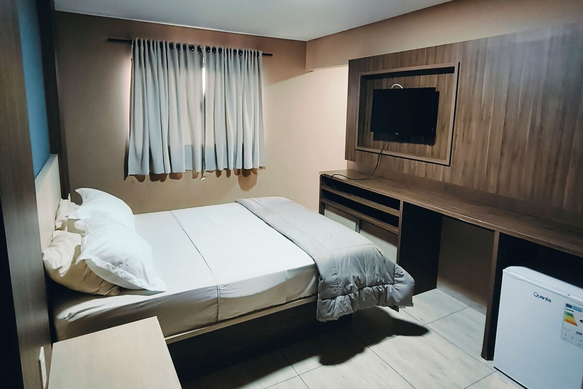 A compact room with a bed, a television mounted on the wall, a small refrigerator, and a curtain.