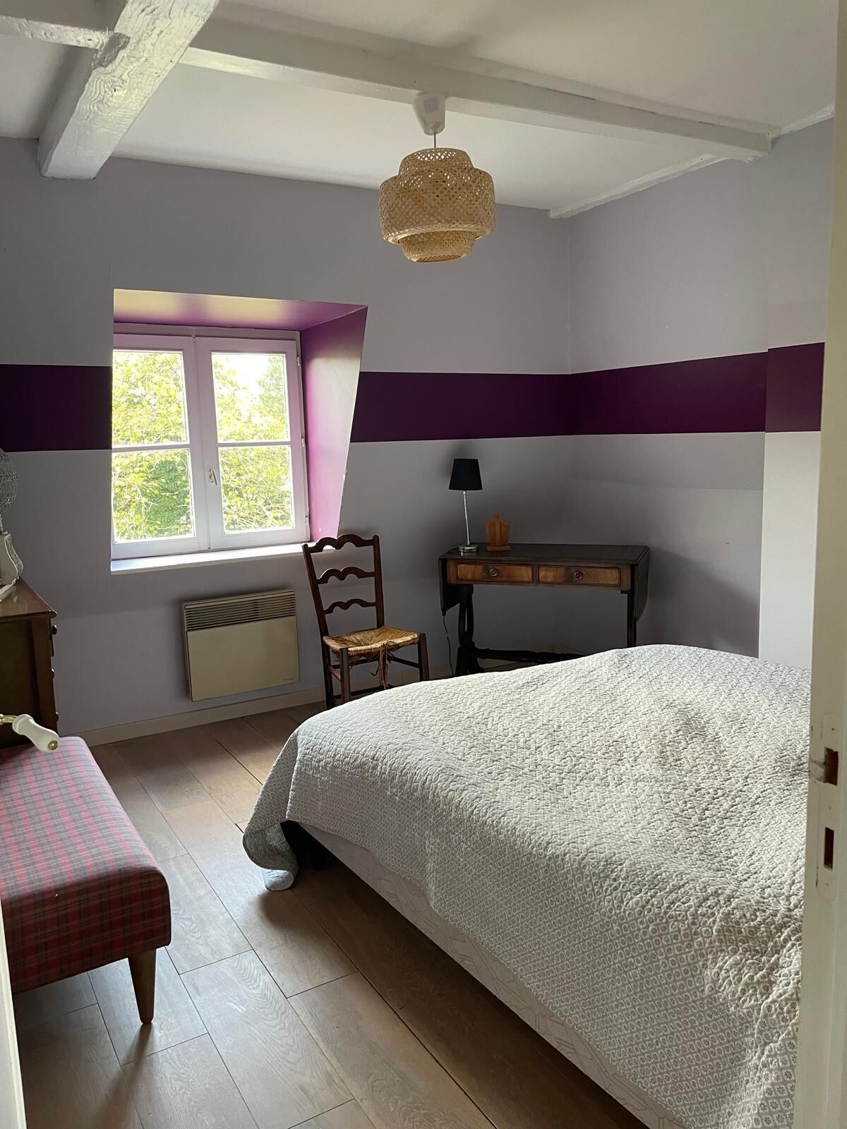 A bedroom with a bed, a chair, a desk, a window, and a hanging light fixture.