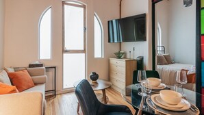 Un espace de vie compact comprenant un canapé, une table à manger pour deux personnes, un téléviseur fixé au mur et un lit avec une table de chevet.