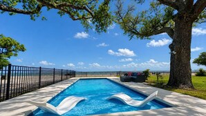 Ein rechteckiger Swimmingpool mit zwei weißen Liegestühlen, umgeben von einem schwarzen Zaun, mit einem Baum und blauem Himmel im Hintergrund.