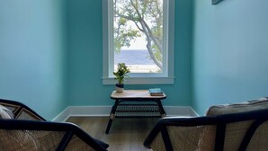 Ein Zimmer mit türkisfarbenen Wänden, einem Fenster mit Meerblick, einem kleinen Tisch mit Pflanze und Korbmöbeln.