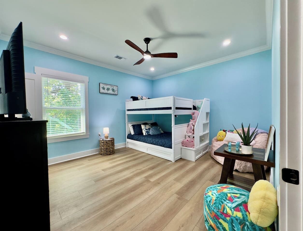 A room with a bunk bed, a ceiling fan, a television, a window with blinds, and a small table with a plant.