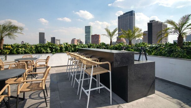 A rooftop bar with wicker chairs and tables, a black counter, and a city skyline view. - City-Core Loft w/ Terrace + Rooftop Views (Mexico City)