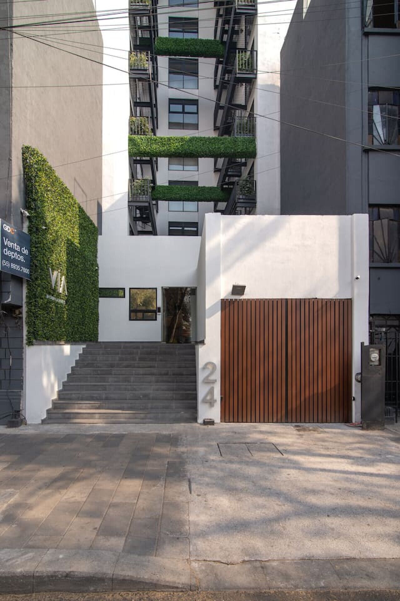 Un bâtiment moderne doté d’un escalier menant à une entrée fermée par un portail, d’un jardin vertical verdoyant et d’une enseigne indiquant une vente.
