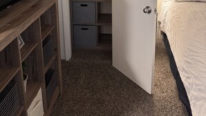 Quarto com cama, televisão sobre um suporte de madeira, armário com cesto de roupa e porta para outro cômodo.