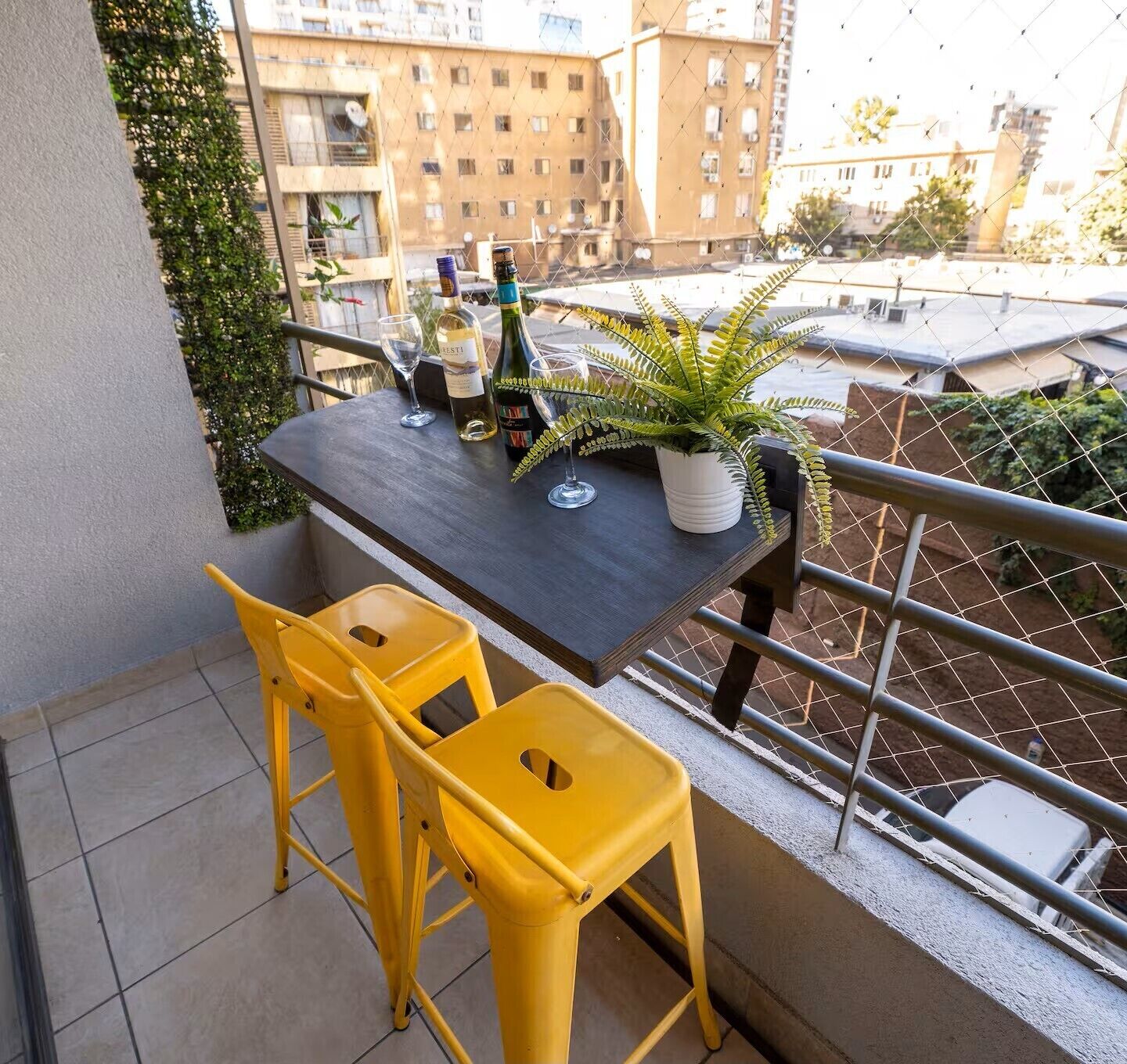A balcony with a table, two yellow chairs, a potted plant, and bottles of wine.
