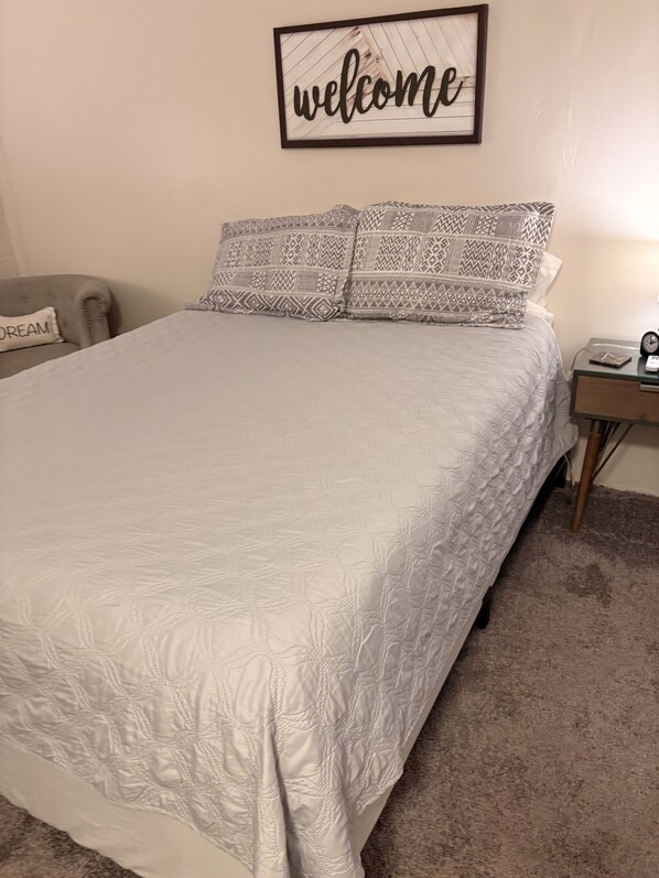 Een bed met een geborgd quilt, een bord met de tekst 'Welkom', een nachtkastje met een klok en een kussen met de tekst 'DREAM'.