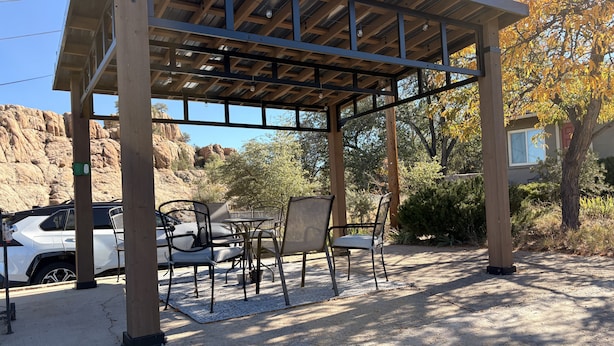 Un'area relax all'aperto, riparata, con tavoli e sedie, un veicolo bianco nelle vicinanze e uno sfondo naturale con alberi e rocce.