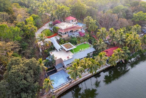 An aerial view of a property with a swimming pool, a tennis court, and a red-roofed building surrounded by dense tropical vegetation.