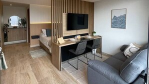 Una habitación de hotel moderna con una cama, un escritorio con televisor, un sofá y una cocina al fondo.