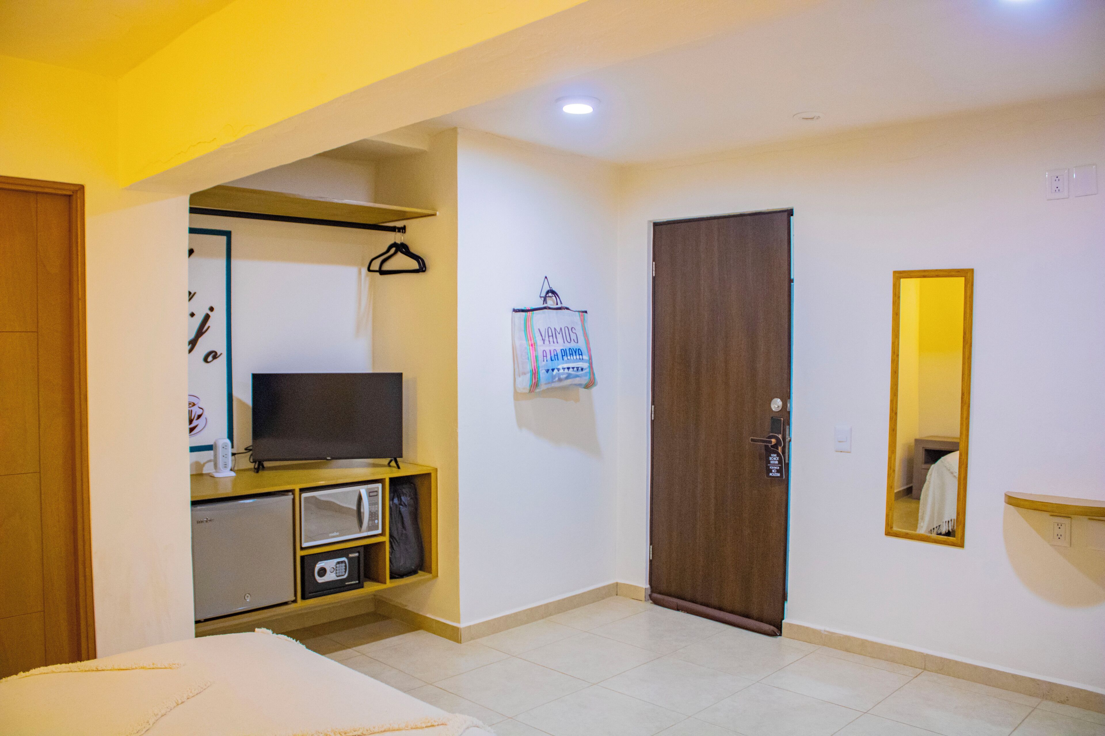 Une chambre d’hôtel avec un lit, un téléviseur posé sur une commode en bois, un four à micro-ondes, un petit réfrigérateur, une porte en bois et un miroir au mur.