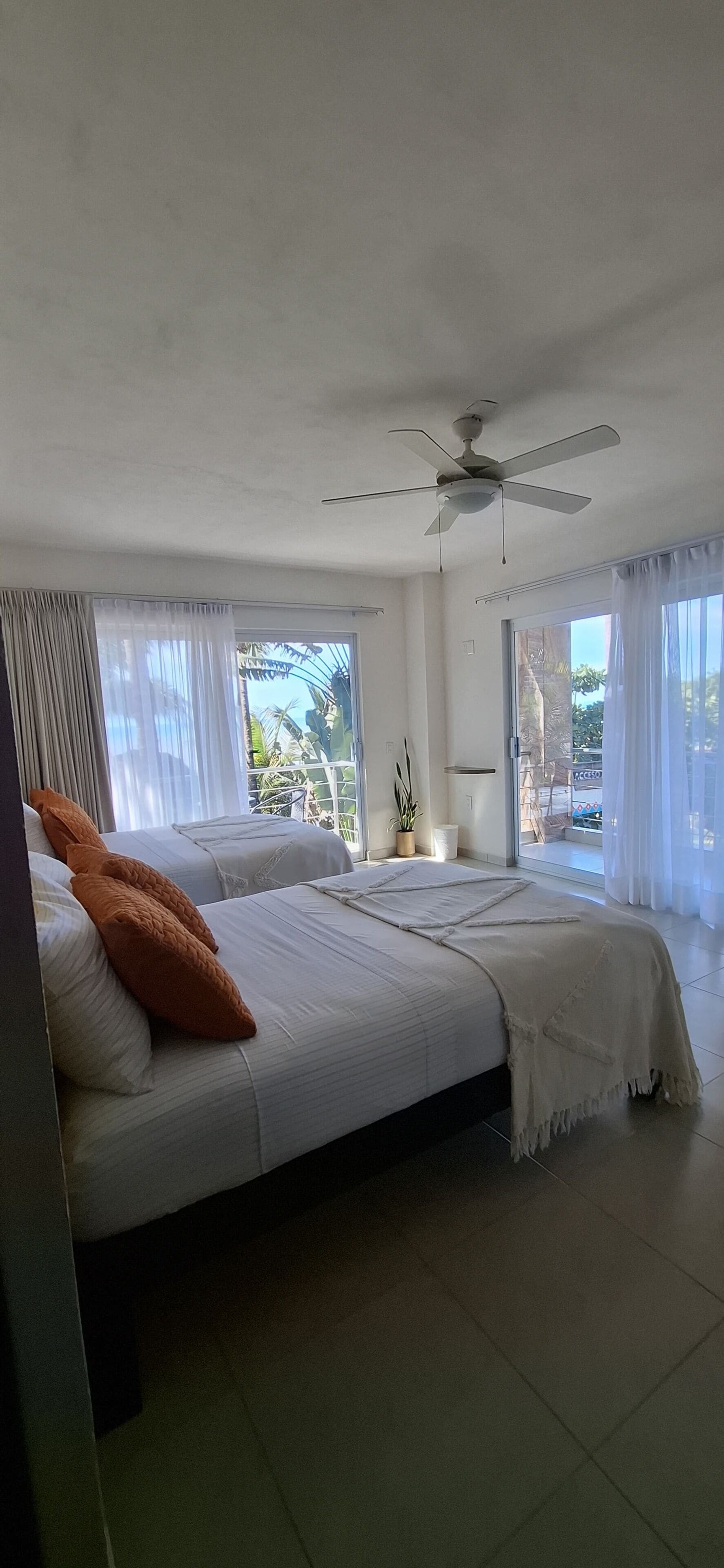Ein Hotelzimmer mit zwei Betten, einem Deckenventilator, großen Fenstern mit Vorhängen und Blick ins Freie.