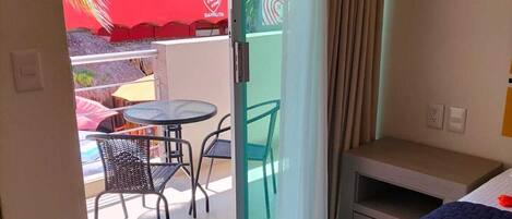 A hotel room with a bed, a small table, a chair, and a sliding glass door leading to an outdoor area with a table and chairs.
