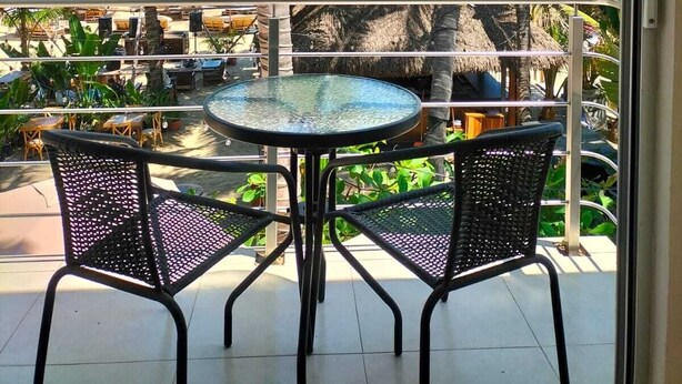 Een balkon met een glazen tafel en twee stoelen, met uitzicht op een strand met palmbomen en een hut met rieten dak.