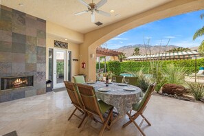 A dining area with a round table, chairs, a fireplace, and a view of a pool and mountains. - Private Palm Springs Retreat w/ Pool, Spa, Mountain Views & Secluded Outdoor Living (Palm Springs)