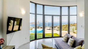 A modern living room with a large window, a flat-screen TV, and a grey sofa with patterned cushions. - Seaview Villa Private Pool & BBQ, 1km from Elounda (Schisma Eloundas)