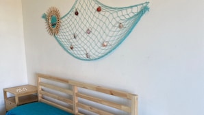 Un dormitorio con una cama, una mesita de noche de madera, una pequeña mesa de madera y un adorno de conchas marinas en la pared.