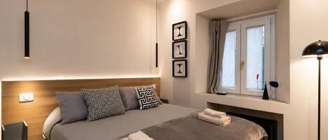 A modern bedroom with a large bed, bedside tables, a window with curtains, and wall-mounted artwork.