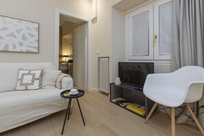 A modern living room with a sofa, a TV stand, a white chair, and a small table. - One-bedroom apartment on the first floor in the heart of Brera, Milan’s artistic and bohemian district. A strategic location just steps from the Pinacoteca di Brera, La Scala Theatre, and Castello Sforzesco.An elegant apartment offering all modern comf (Milan)