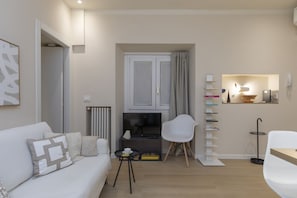 Ein modernes Wohnzimmer mit einem weißen Sofa, einem Fernseher, einem weißen Sessel, einem kleinen Tisch und einem Bücherregal.