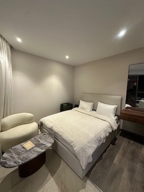 A modern bedroom with a bed, a chair, a small table, and a nightstand.