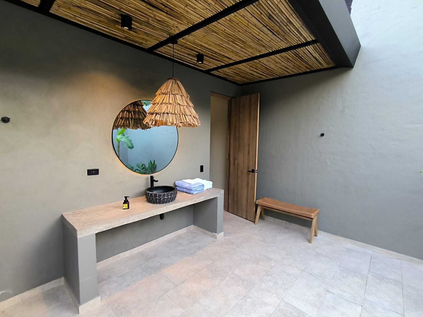 A bathroom with a round mirror, a black vessel sink, a wooden bench, and a ceiling with a woven design.