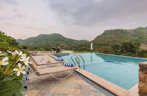 A pool with lounge chairs and a view of hills. - Brijbagh by ORUVilla (Kumawaton Ka Gurha)