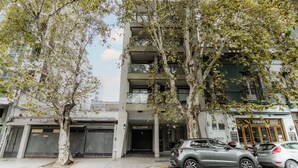 Um edifício com vários andares, entrada central, área de estacionamento com carros e árvores na frente.