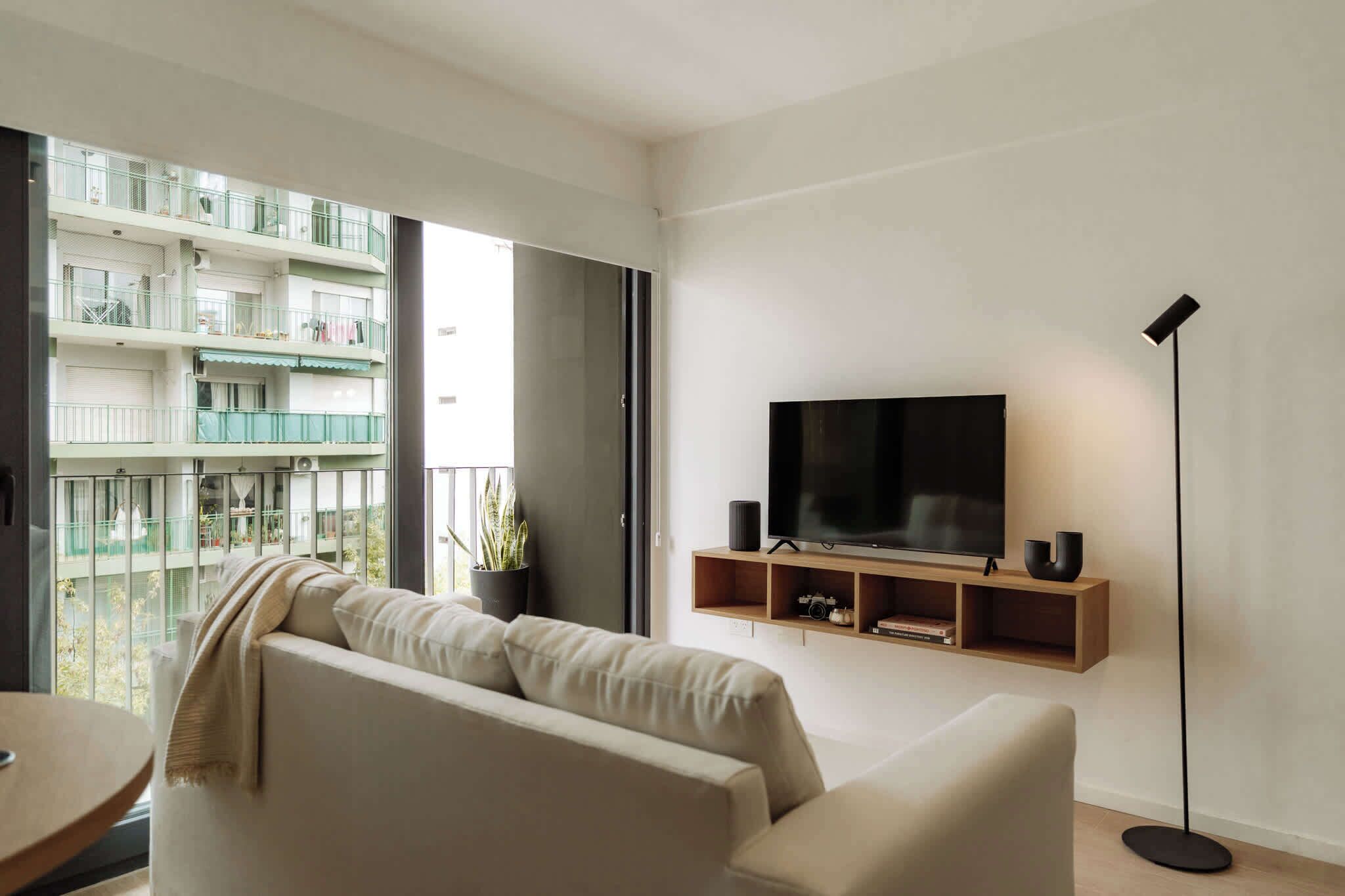 Una sala de estar moderna con sofá, televisor montado en la pared, lámpara de pie y balcón con vista a otros edificios.