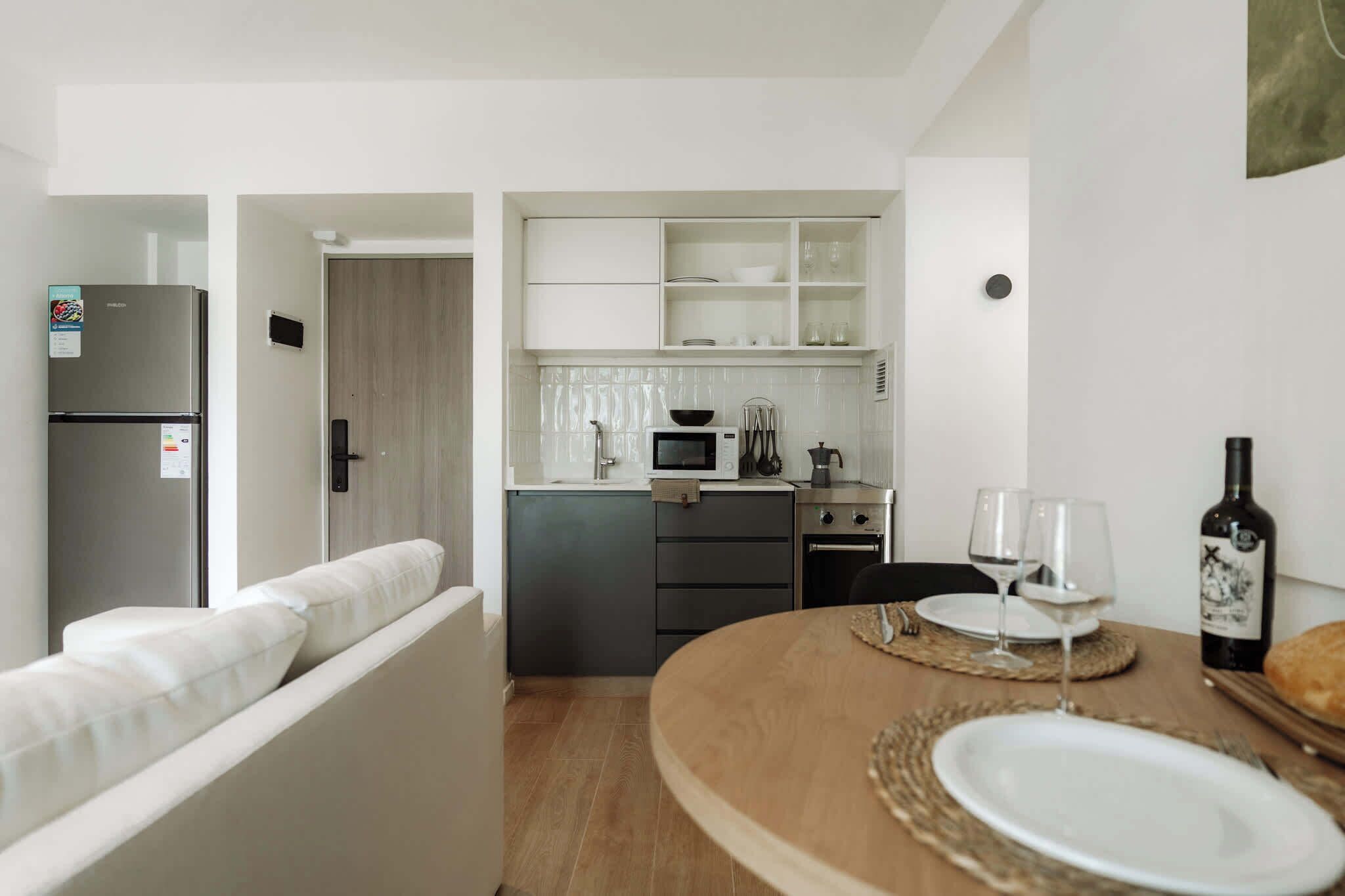 A compact kitchen with a refrigerator, microwave, and oven. There is a dining table with plates and glasses, and a bottle of wine.
