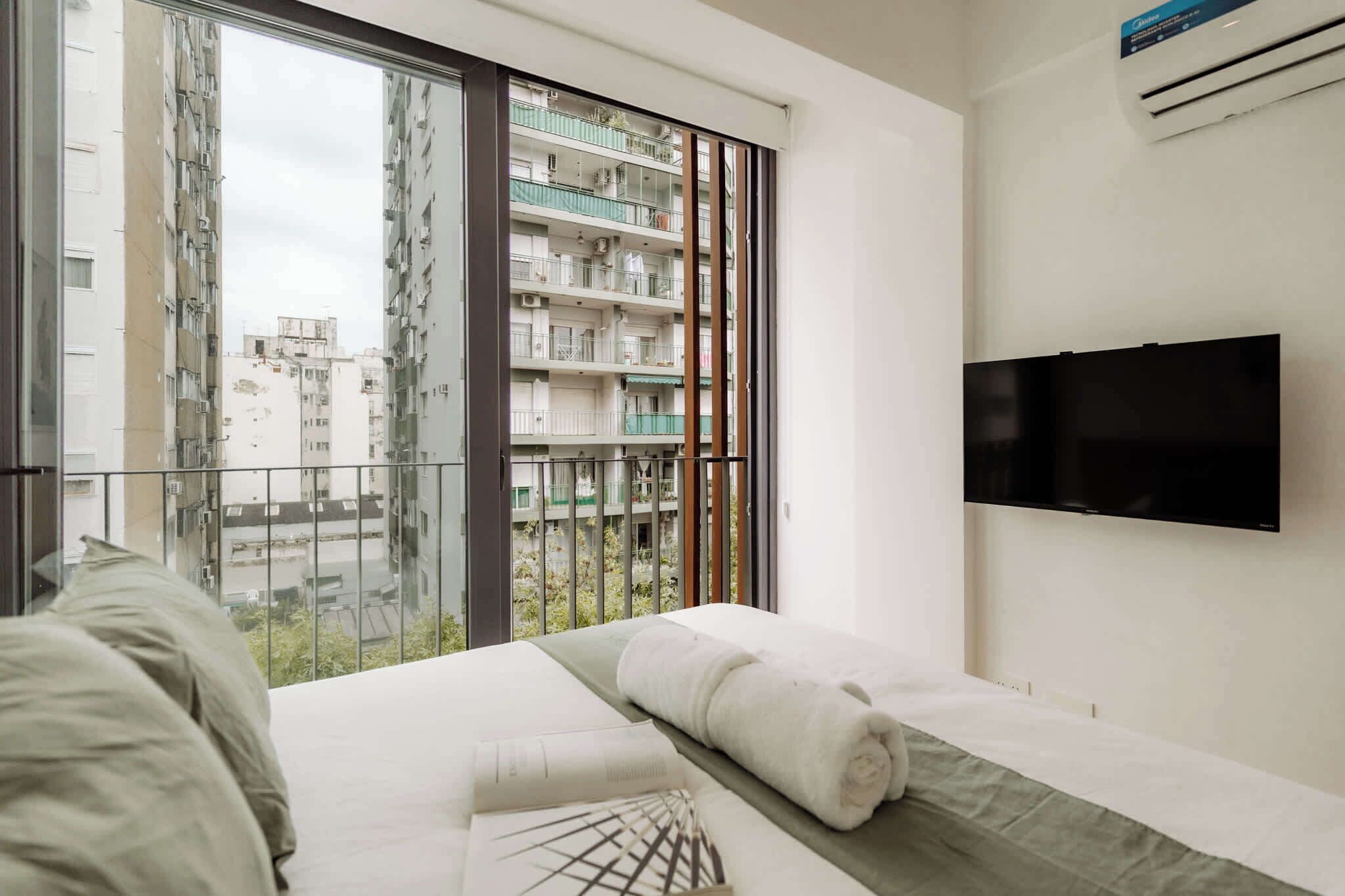 Habitación de hotel con un ventanal grande, una cama con colchón y almohadas, un televisor de pantalla plana montado en la pared y un equipo de aire acondicionado.