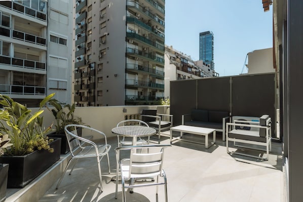 Terraço no telhado com mobiliário para áreas externas, plantas em vasos e vista para edifícios altos ao redor.
