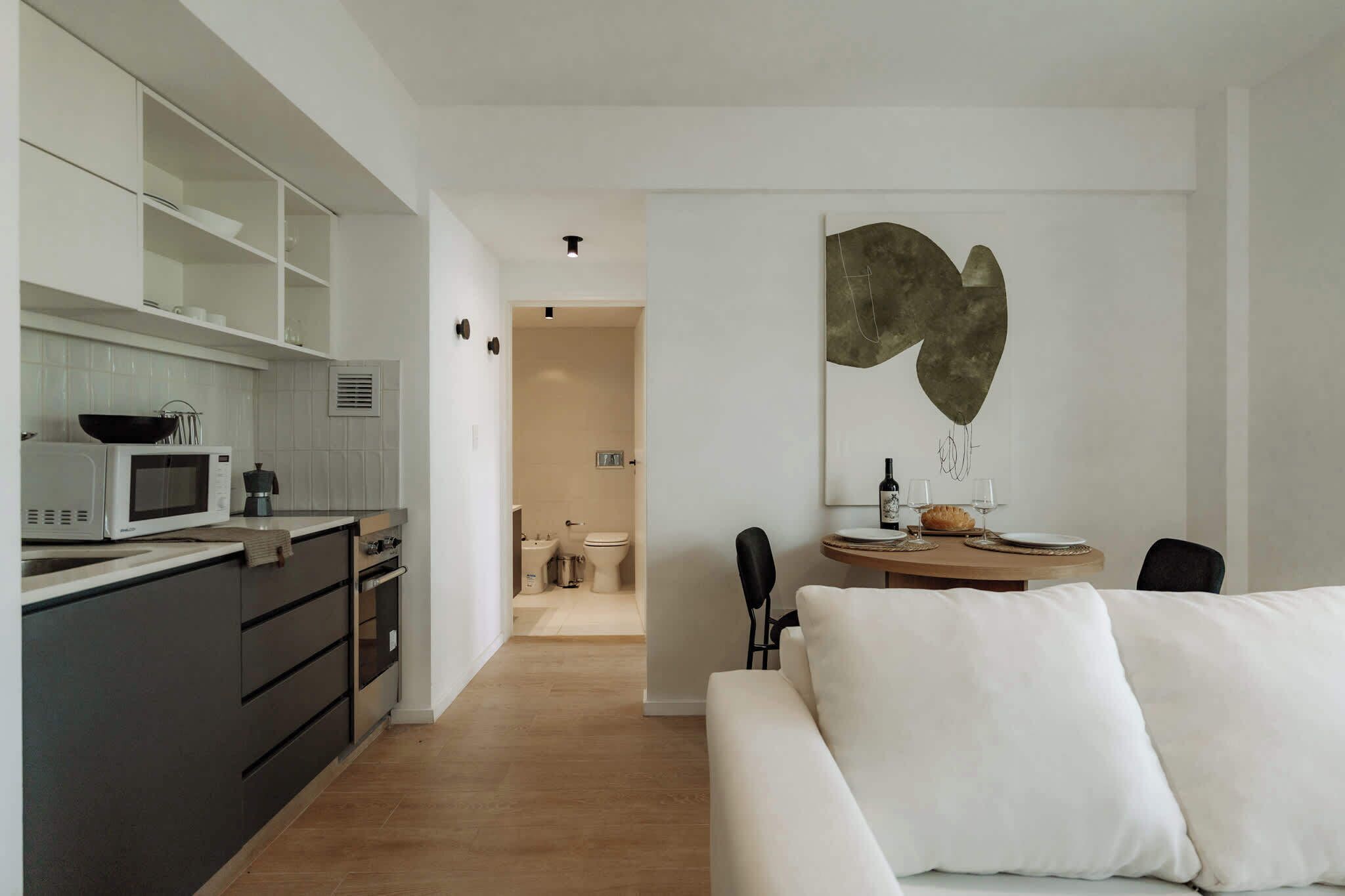 Una cocina moderna con armarios blancos, un microondas y un lavamanos. Hay una zona de comedor con mesa y sillas, y un sofá blanco en primer plano.