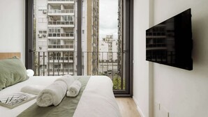Quarto de hotel com uma cama, um cobertor verde, duas toalhas enroladas, uma janela com vista para edifícios e uma televisão na parede.