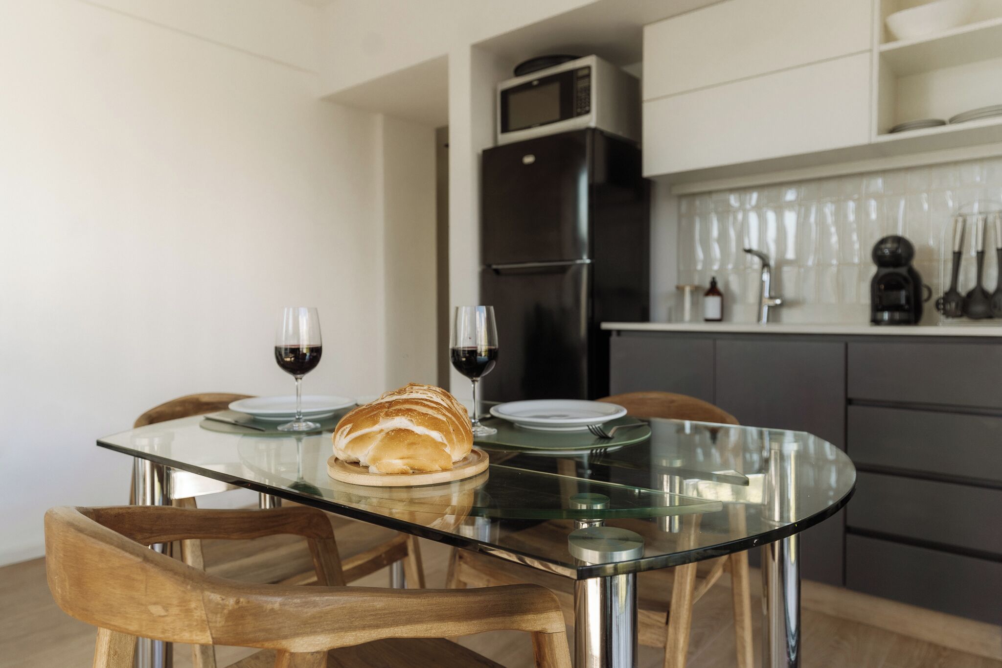 Una cocina moderna con una mesa de comedor de vidrio, sillas de madera, una hogaza de pan y dos copas de vino tinto.