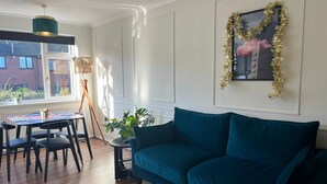 A modern living room with a teal sofa, a dining table with chairs, a small side table with a plant, and a framed picture with a gold tinsel garland. - Modern Classic 2 Bed Apartment Near Graves Park (Sheffield)