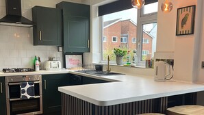 A modern kitchen with a central island, bar stools, a trash bin, and a view of a brick building. - Modern Classic 2 Bed Apartment Near Graves Park (Sheffield)