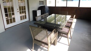 Un patio coperto con tavolo in vetro e sedie in vimini, un barbecue e una porta bianca con pannelli di vetro.